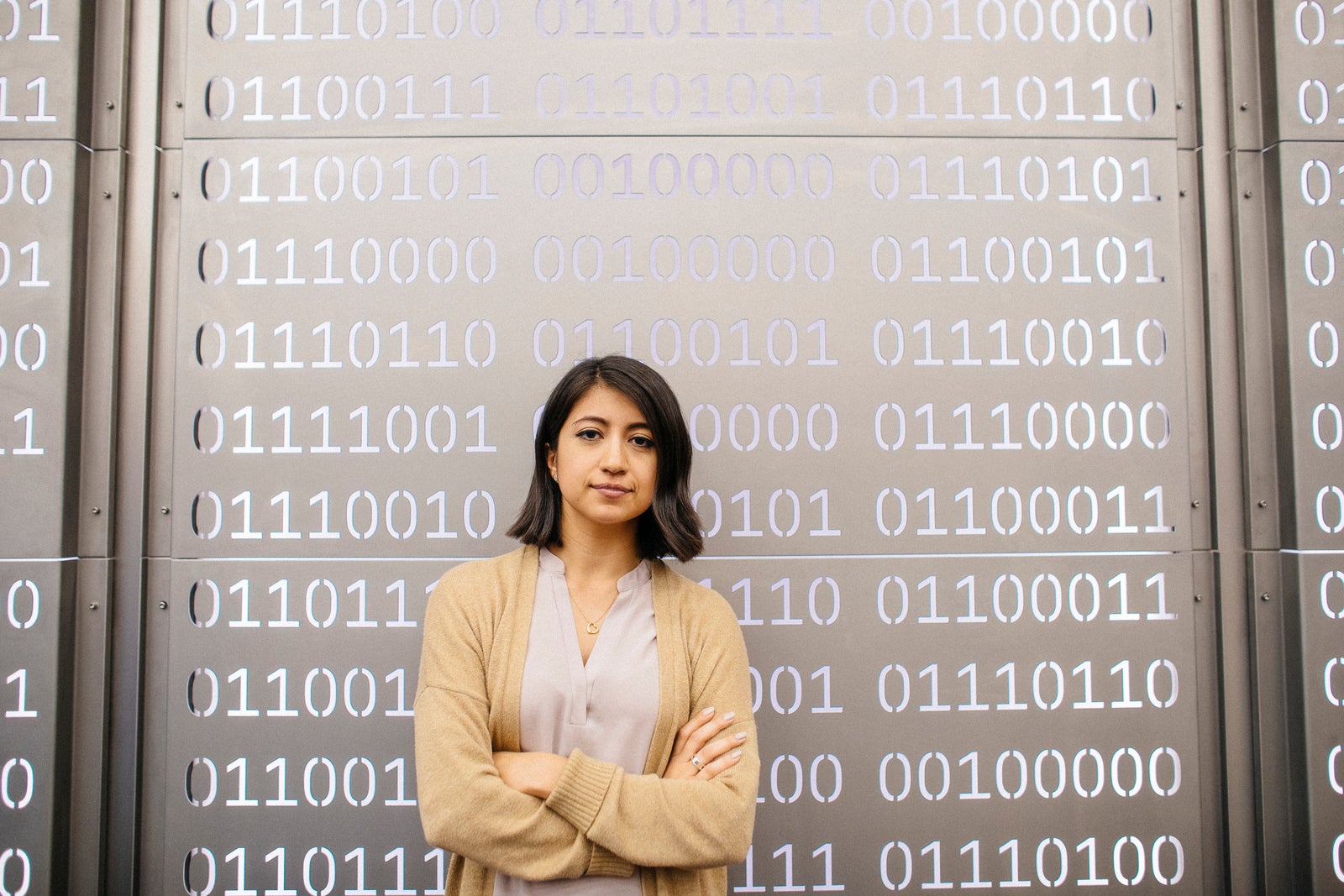 Woman crosses arms over chest and looks at camera, against a metal wall with punched out 0s and 1s, meant to represent computer code