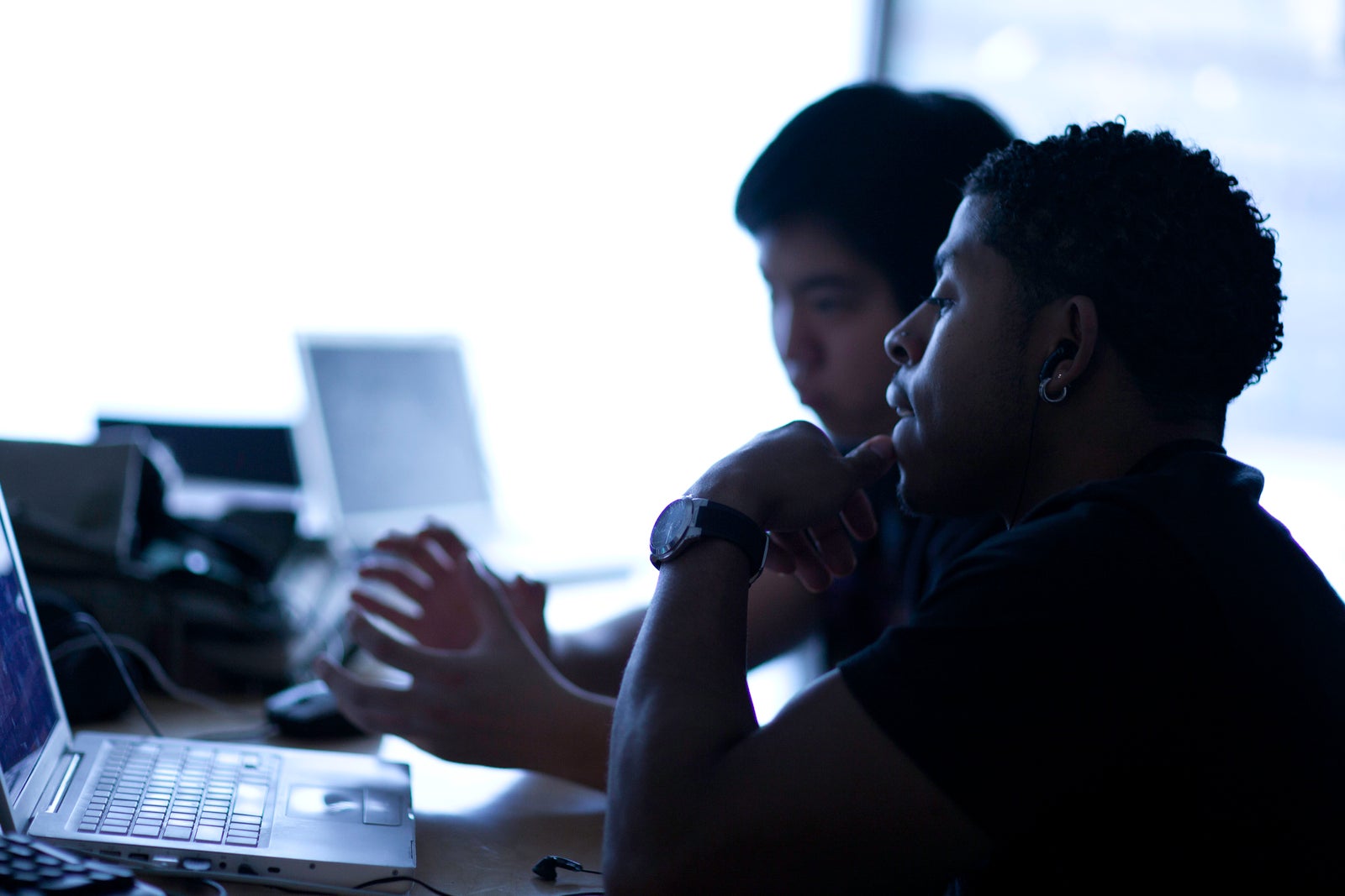 Two male college students type on laptops. Their profiles are visible but they're mostly backlit.