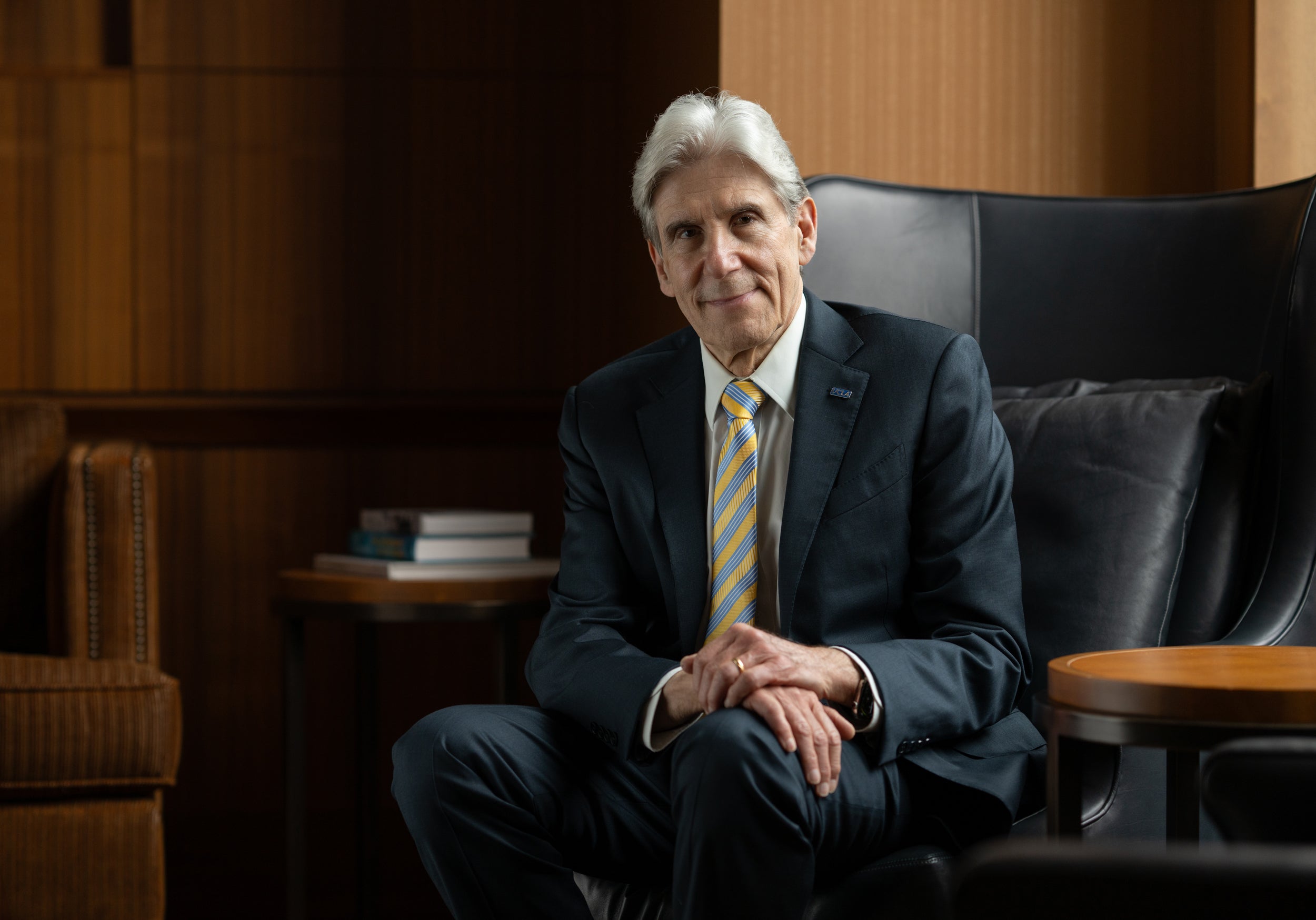 Man with white hair and a blue and gold tie sits in a wood-paneled office on a leather chair