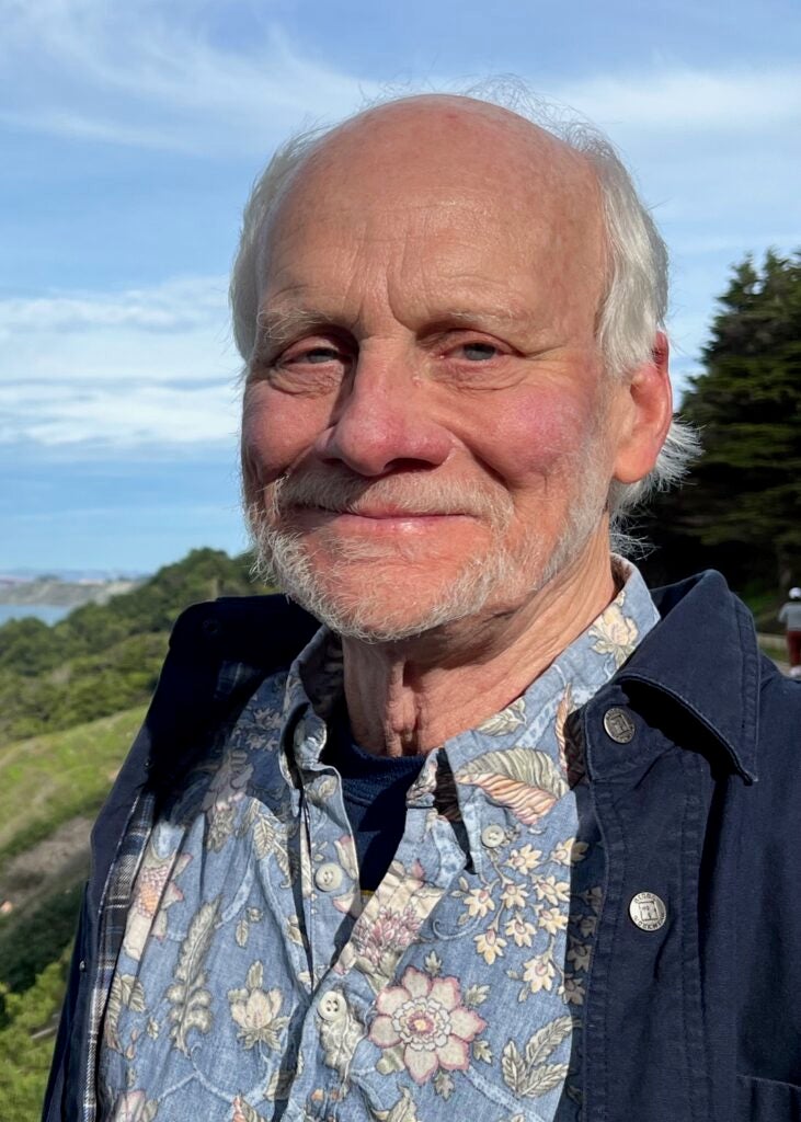 Kent Lightfood smiles at the camera wearing a floral shirt and jacket, outside on a green hillside