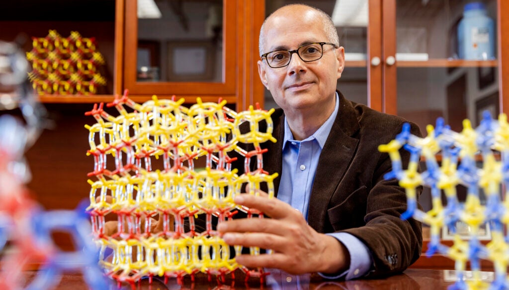 Man in tweed jacket in an office lined with wooden cabinets, holding a large model of a molecule assembled with smaller parts