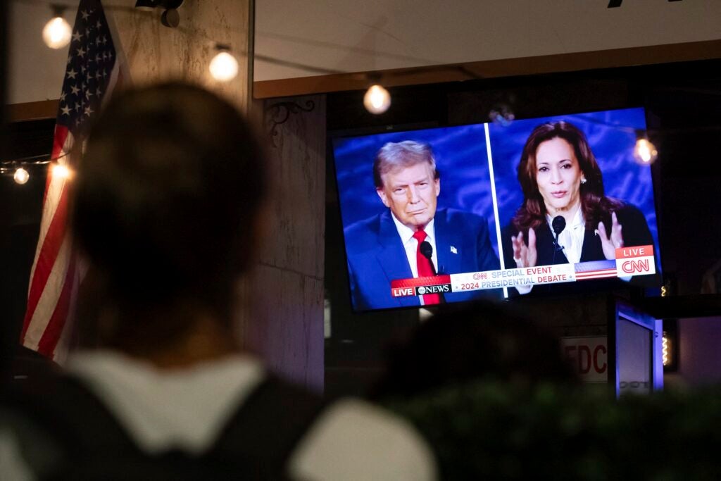 A split-screen image on a TV shows Donald Trump and Kamala Harris debating, being watched by people at a bar