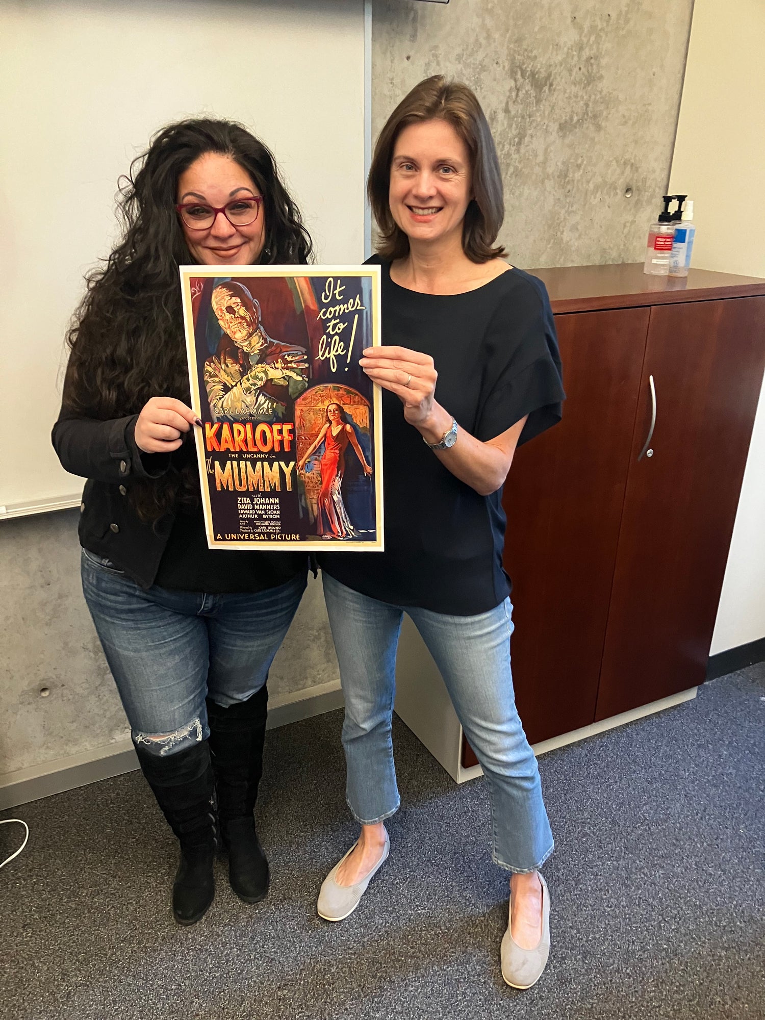 Two smiling women hold a poster of the old movie The Mummy in a classroom