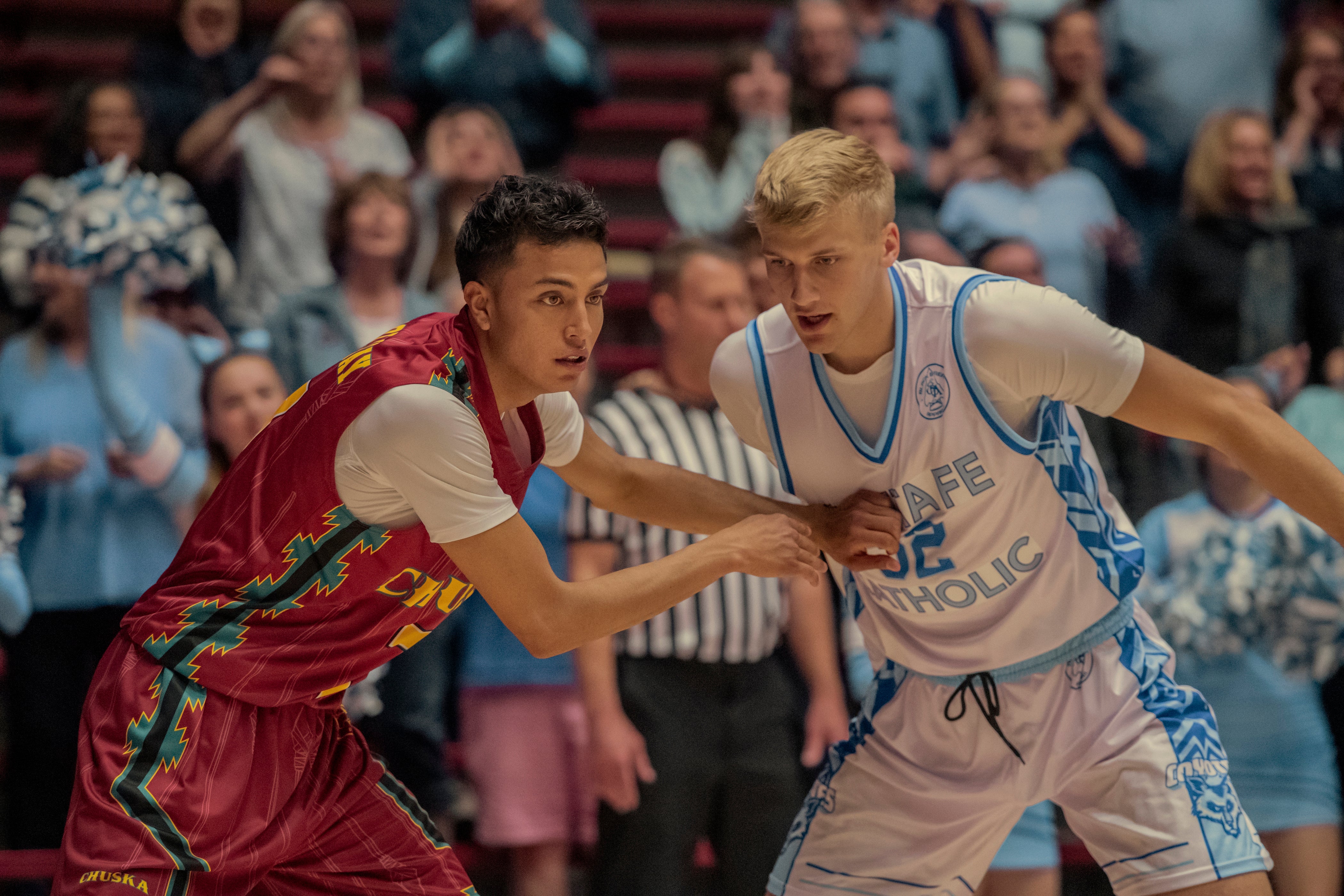A young man with dark hair in a red basketball jersey and a young man with blonde hair in a white basketball jersey play in front of a crowd