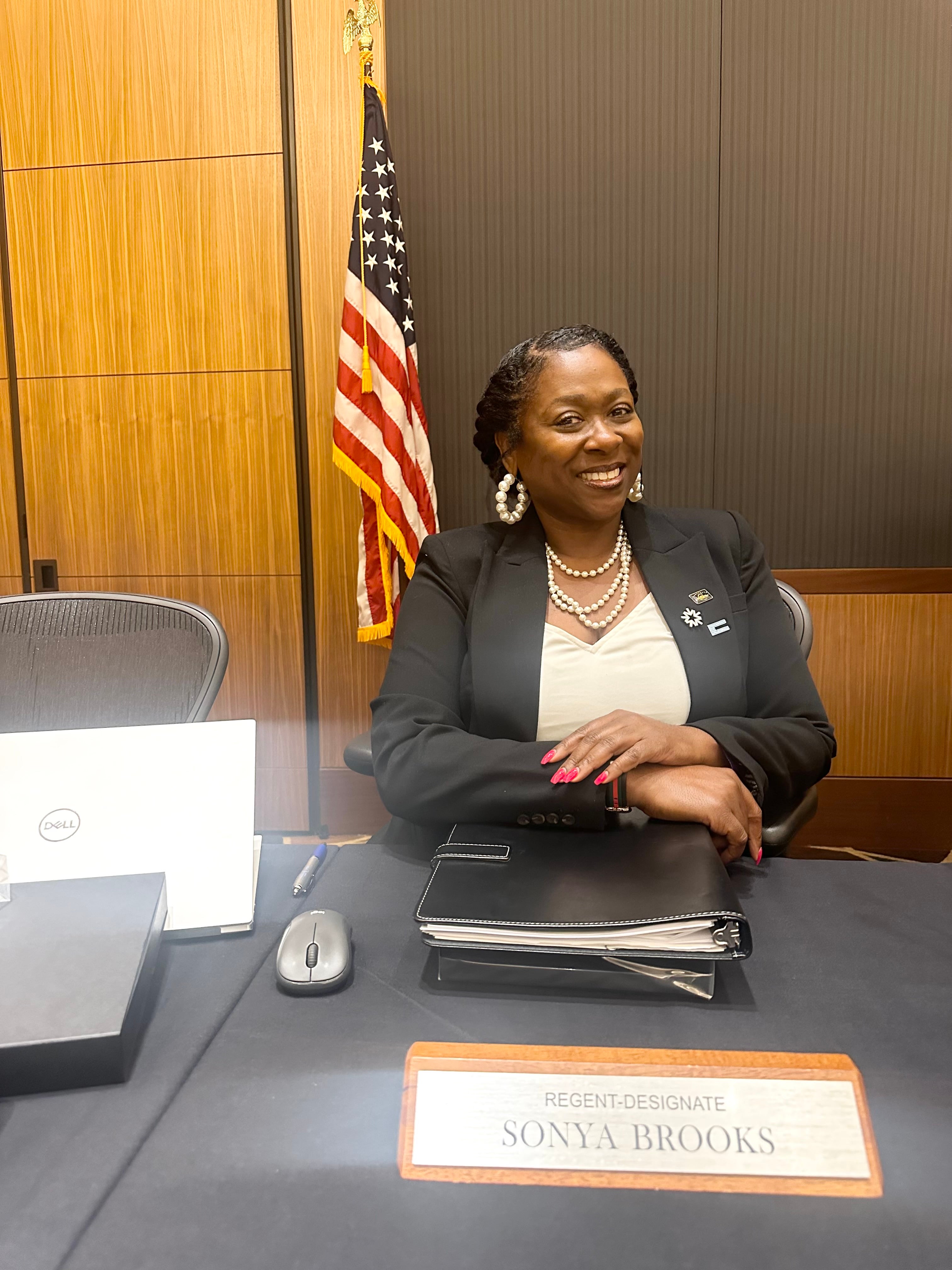 Sonya Brooks smiles at the camera at her spot around the UC Regent's meeting table, in front of her name placard.