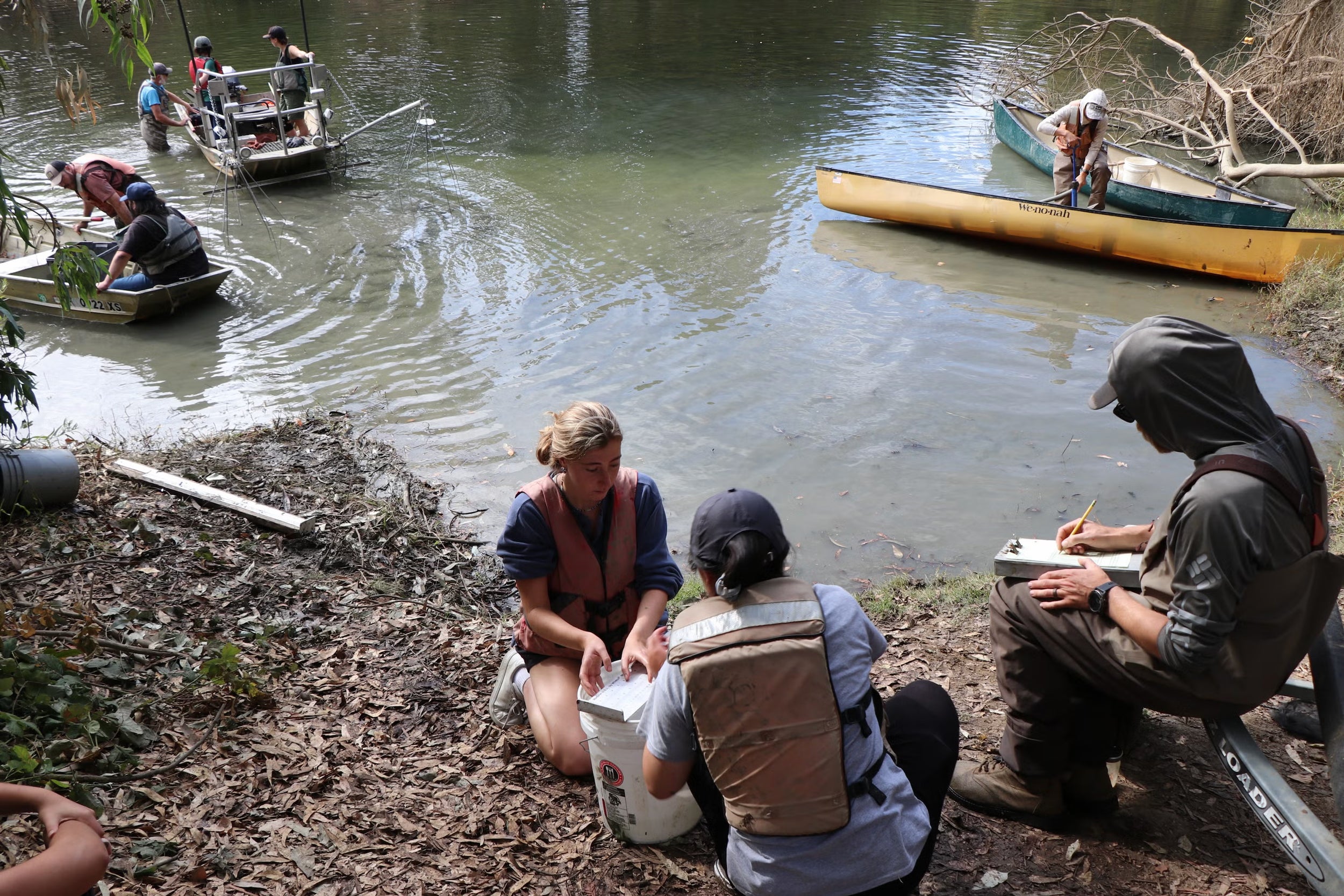 People in boats in a creek and sitting at the edge of the water writing and inspecting equipnment