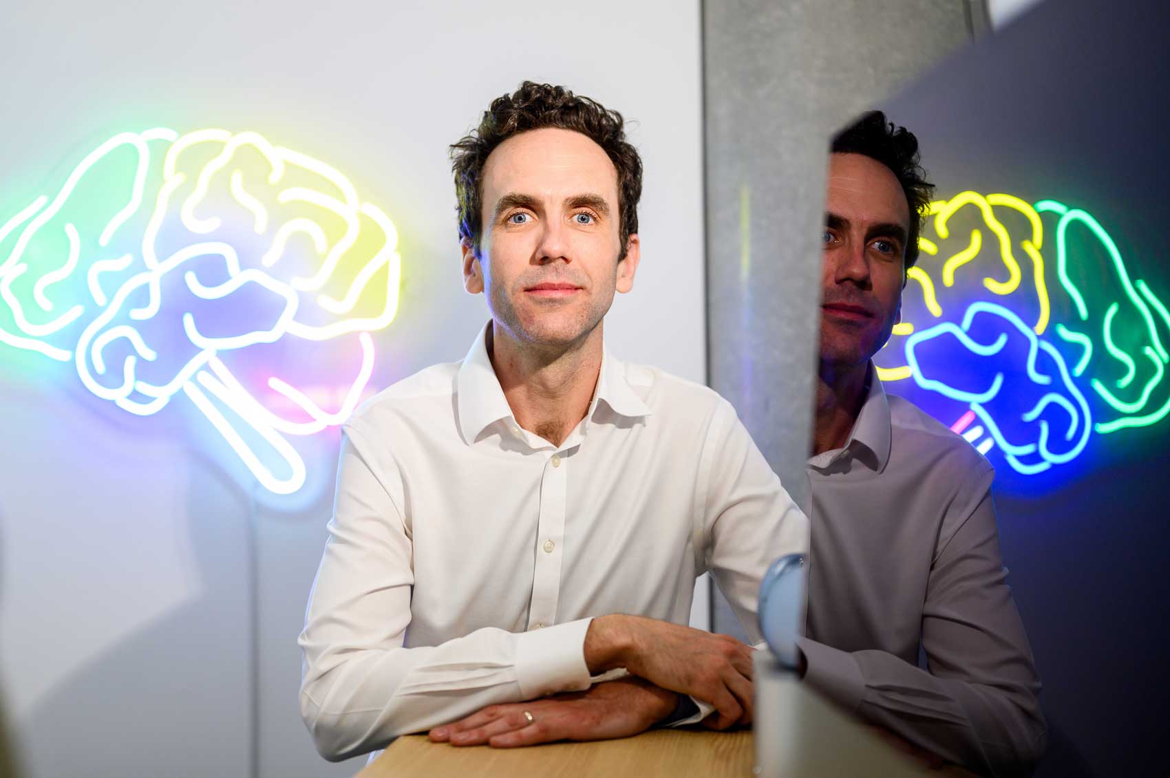 Man in white dress-shirt looks at camera while siting at a desk with a colorfully lit brain illustration lamp behind him