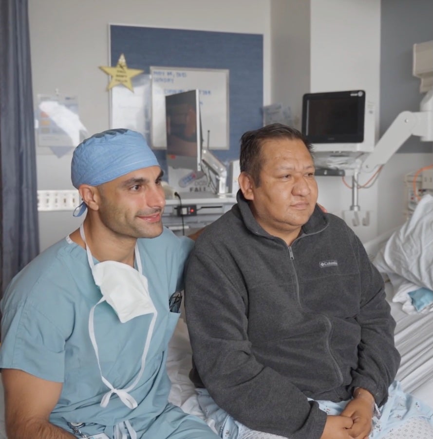 A surgeon in scrubs and a patient sitting up in a hospital bed pose together