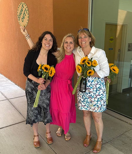 Three women pose together, holding sunflower bouquets