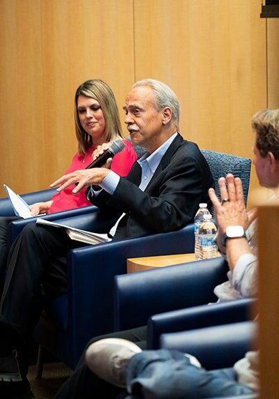 In a wood-paneled room, a seated man, center, gestures as he talks into a microphone. A woman looks on in the chair to his right