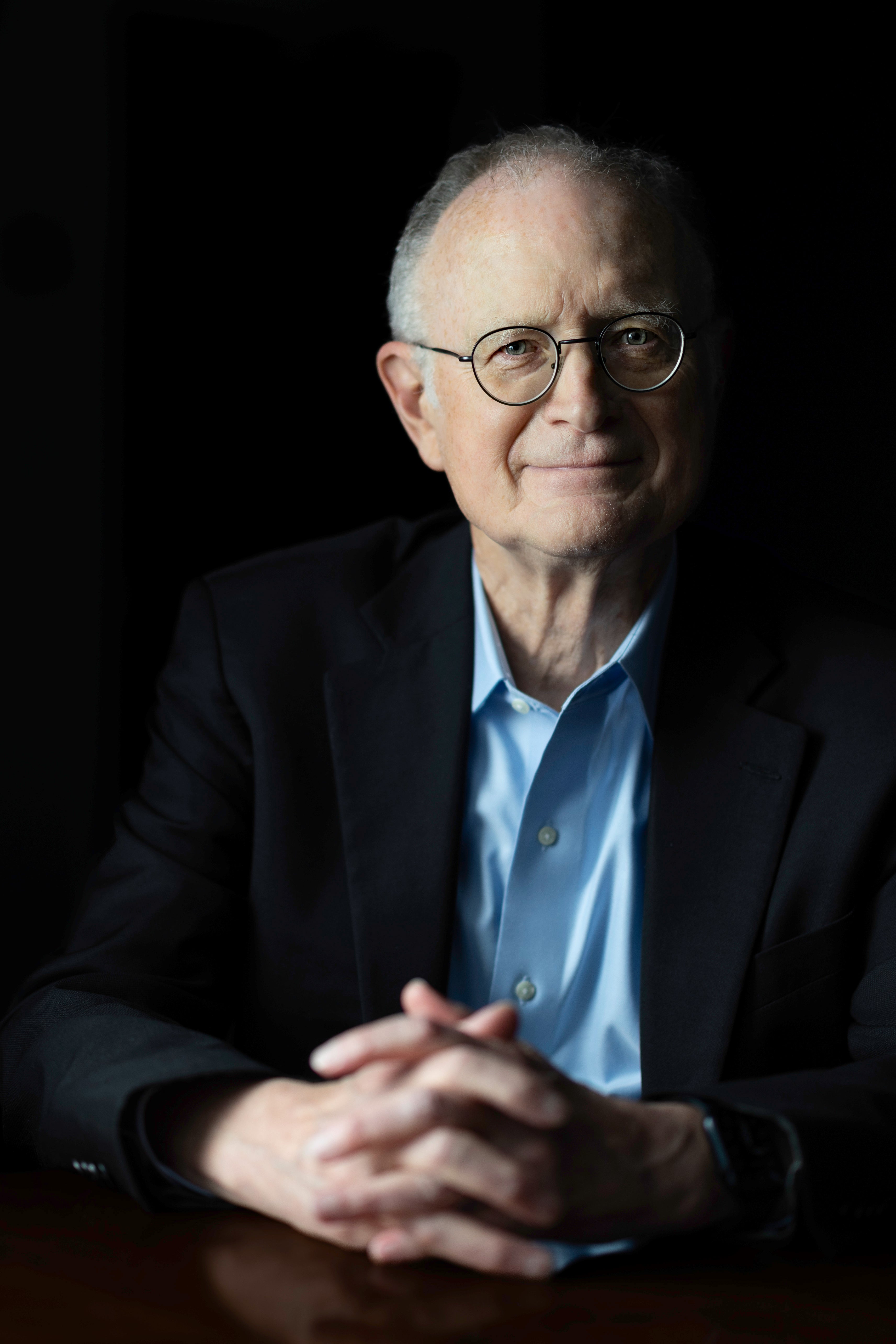A man wearing glasses smiles at the camera, hands folded