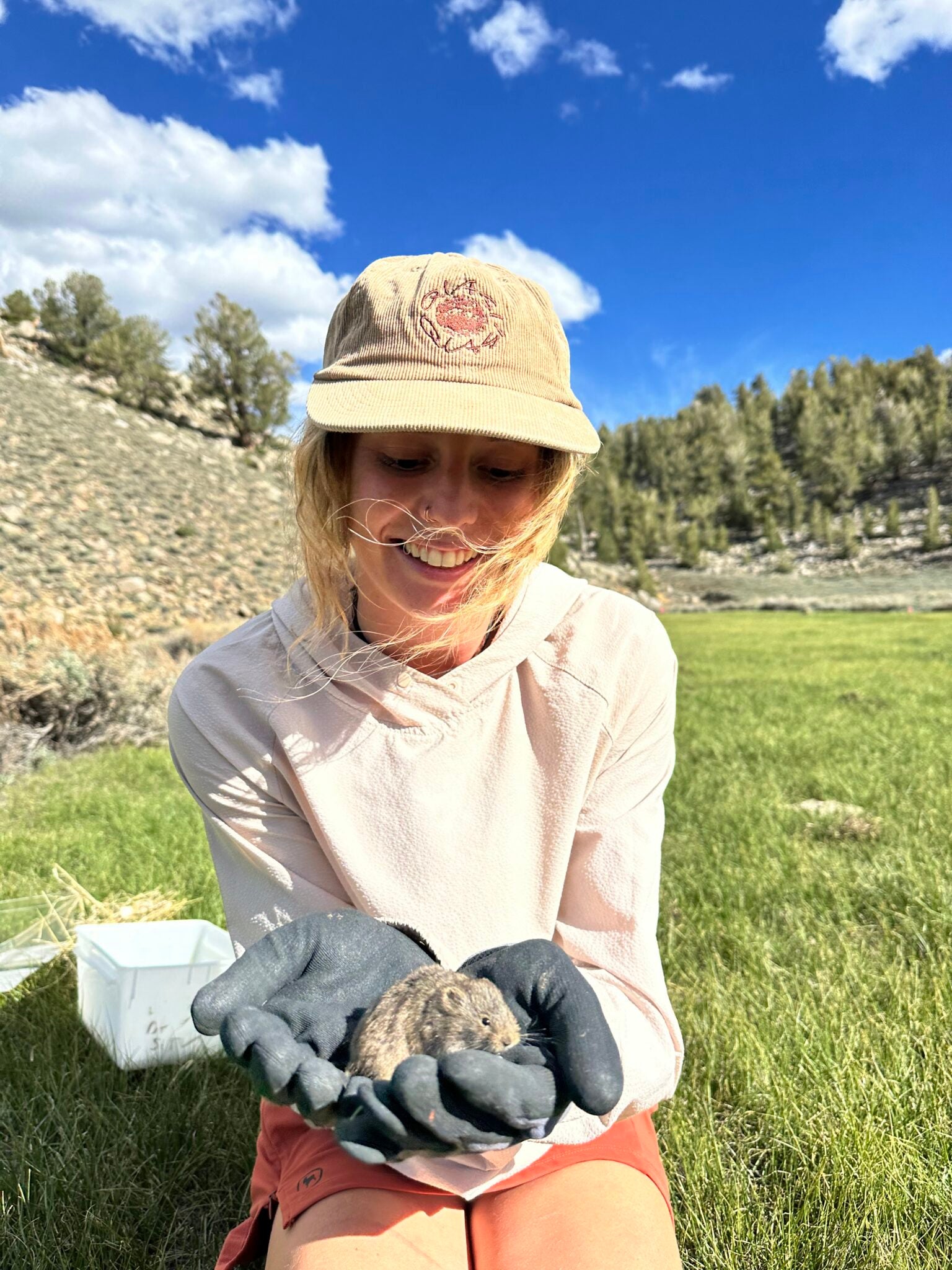 a blond woman in a cap kneeling in a grassy meadow holding a small brown rodent in her gloved hands