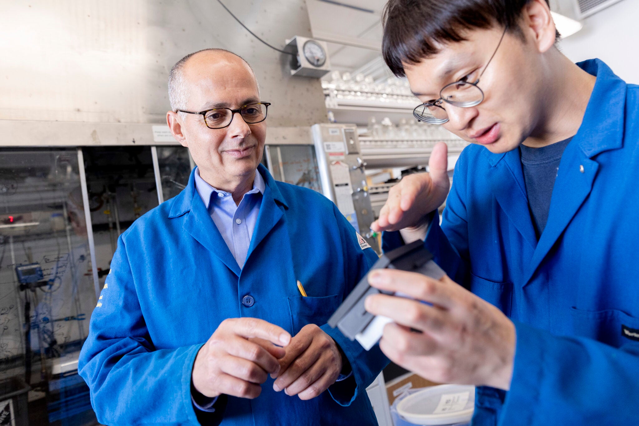 A man in glasses and a blue lab coat watches a young man hold an object and gesture at it