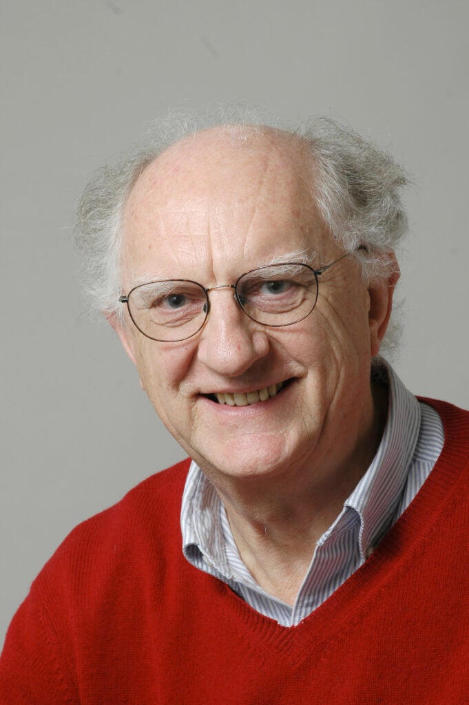 John Clarke smiles at the camera wearing a red sweater.