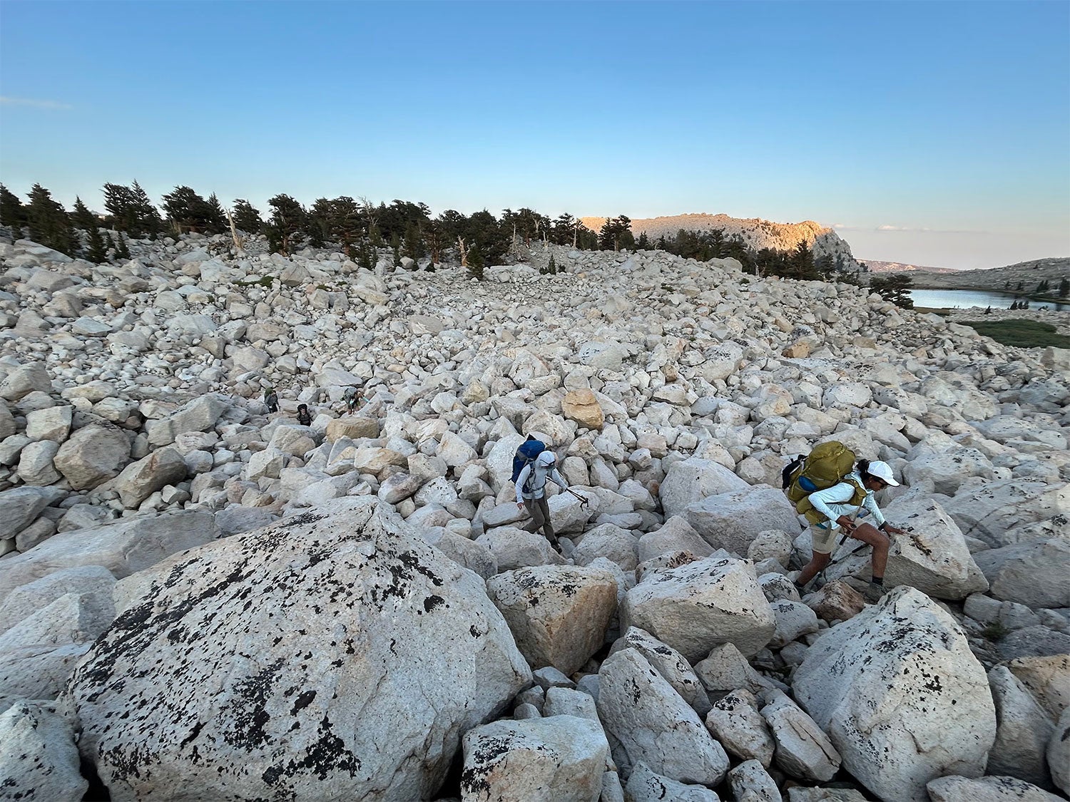 Two hikers in backpacks with poles navigate a granite boulder field.