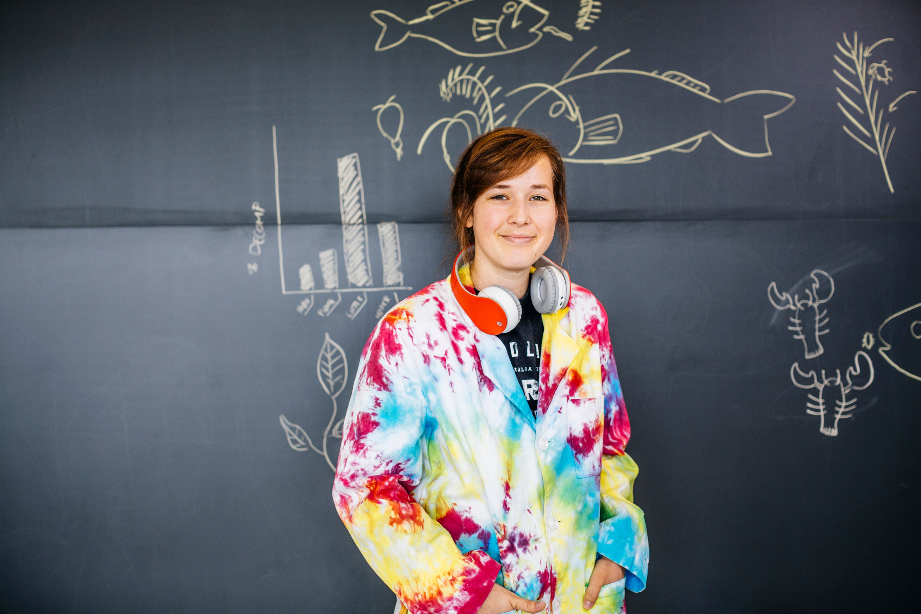 A young woman wearing headphones and a tie-dyed hoodie stands in front of a chalkboard with various drawings on it, including a few fish 