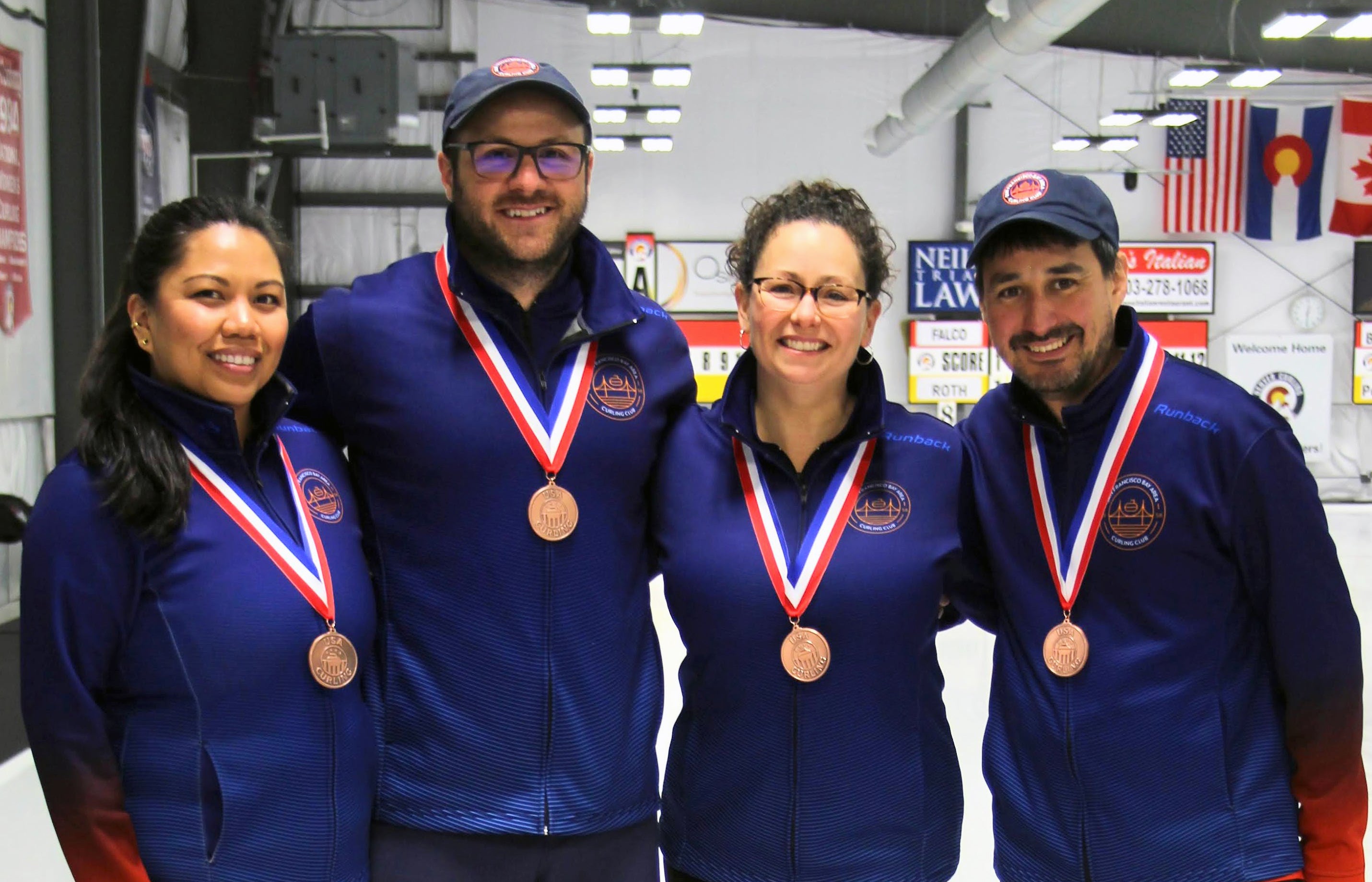 Four people in blue uniforms stand smiling wearing bronze medals