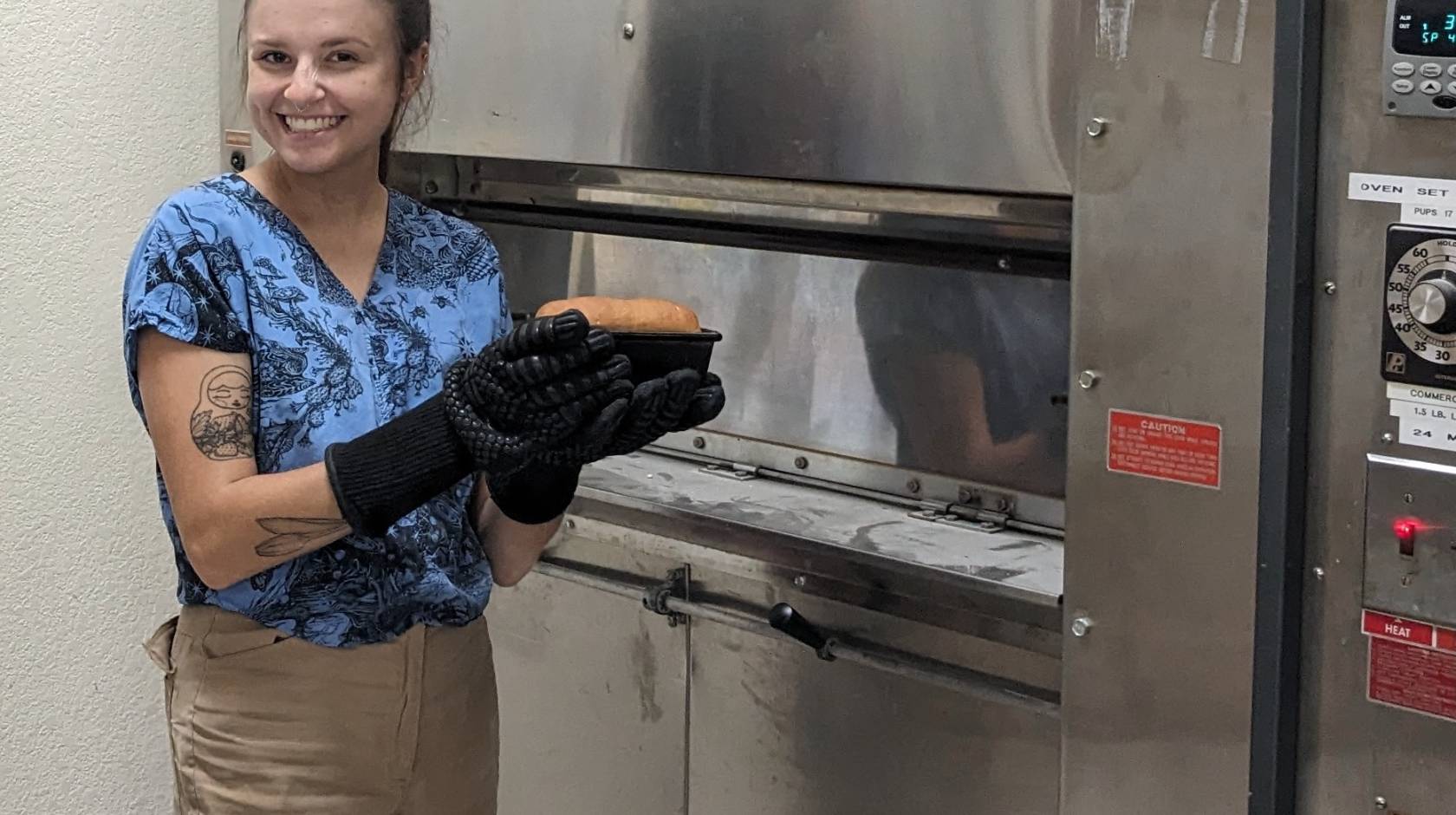 Woman with baking mitts smiles and holds up a loaf of bread in front of an oven