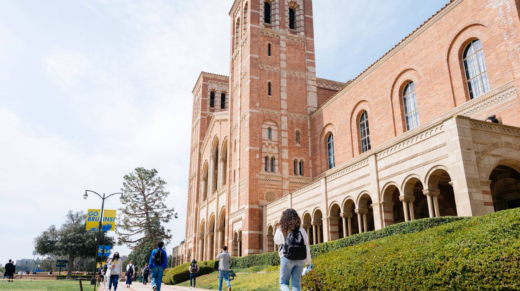 Royce Hall on the UCLA campus, a light adobe brick building, photographed from a low angle and at the side as a few students walk