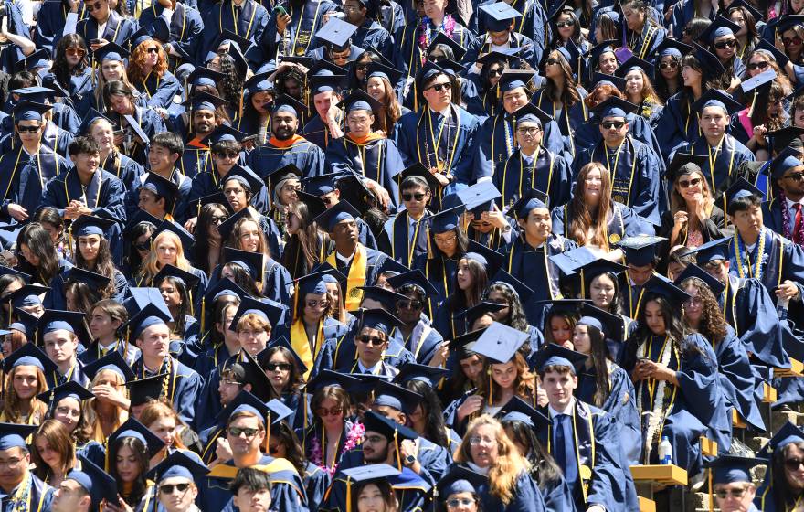 A high-angle shot of a dense crowd of university graduates in blue caps and gowns filling stadium bleachers, accented by colorful leis, stoles and sunglasses under bright sunlight.