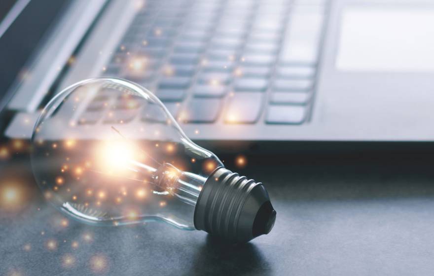 A lit lightbulb laying on its side on a desktop next to an open laptop. Sparkles shimmer around the lightbulb.