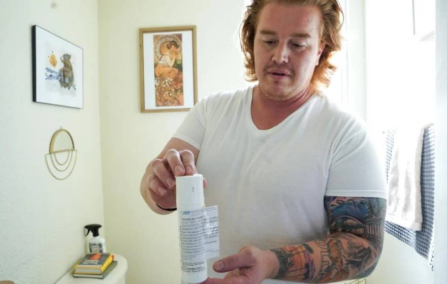 A man with arm tattoos in a white t-shirt holds a white plastic bottle, standing in a well lit room with white walls.