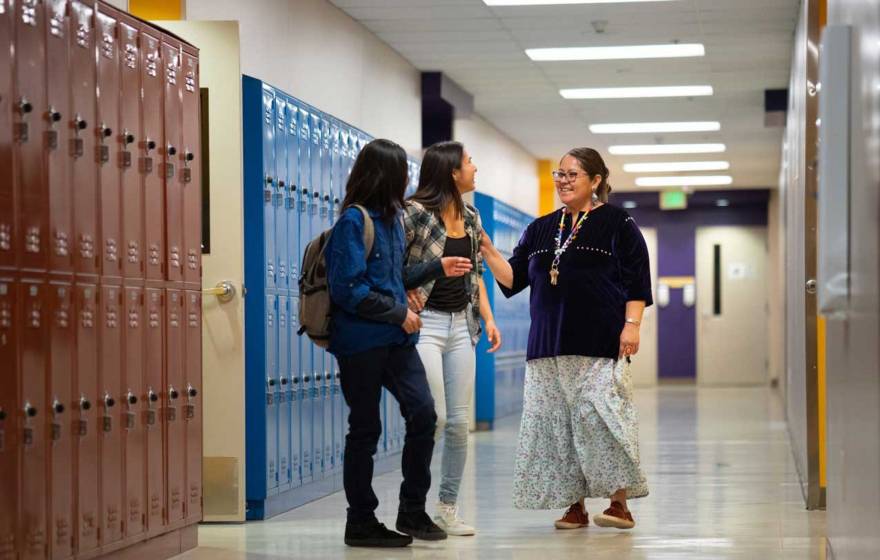 An adult talking with two high school students in a school hallway, with rows of lockers in the background