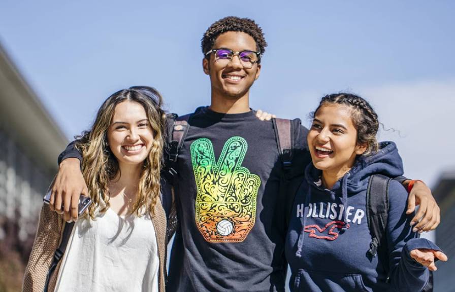 Three young adults smile together outdoors with their arms around each other.