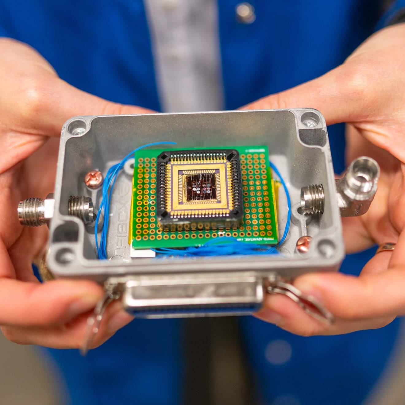 A pair of hands holds a small plastic box that contains a complex electronic chip